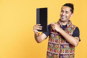 Joyful man wearing traditional indian attire using tablet to take selfies. Ecstatic person with positive facial expressions taking photos using device selfie camera, isolated over studio backdrop