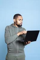 Carefree happy african american guy working on his laptop in studio, wearing a grey hoodie and composing emails to solve his tasks and paperwork. Young adult multitasking, browses websites. photo