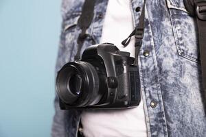 Closeup of a DSLR hanging over an individual wearing a denim jacket and white shirt against an isolated background. Selective focus on a professional camera positioned on the body of a person. photo