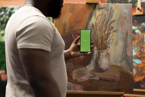 Male artist feeling inspired to paint a new artwork and holding phone with isolated display, thinking about a new painting technique in art and craft atelier. Working with motivation and creativity. photo
