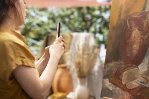 Confident painter presenting her new masterpiece on online branding , filming a vlog on smartphone to promote the artistic products used for the artwork on canvas. Art and craft content creation. photo