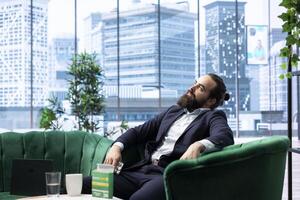 Overworked fatigued boss swallowing an aspirin to ease his headache, taking painkillers to help with his burnout and workload. Young CEO being anxious and overwhelmed in the office. photo