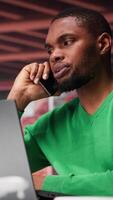 Vertical Freelancer in home office having important phone call with business partner using his mobile phone. African american guy sitting at desk discussing company matters during call. Camera B. video