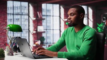 African american remote worker solving his daily freelancing tasks on laptop, working in relaxation from the comfort of his modern loft. Tech savvy black man internet user, multitasking. Camera A. video