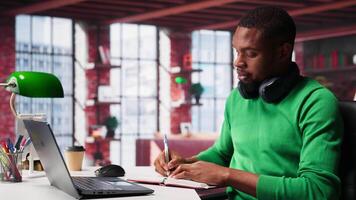 Smiling black man taking notes while watching a tutorial presentation, doing online self learning and writing details. Young adult man writes in his textbook notepad. Camera A. video