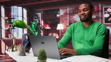 Black remote employee in home office using laptop for web assignments, preparing presentation slides and working on data layouts. Young teleworker finishing company project. Camera B. video