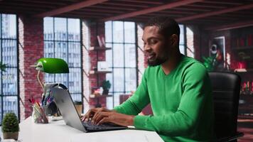 African american remote worker solving his daily freelancing tasks on laptop, working in relaxation from the comfort of his modern loft. Tech savvy black man internet user, multitasking. Camera B. video