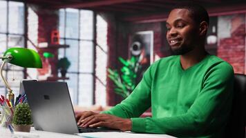 Black remote employee in home office using laptop for web assignments, preparing presentation slides and working on data layouts. Young teleworker finishing company project. Camera A. video