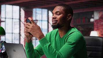 Man at home using smartphone to compose social media posts, producing content as a freelancer. African american guy monitoring profile engagement using mobile phone. Camera A. video