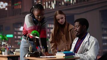 Diverse group of pupils in a library discuss with a medic resident, preparing for a new lecture at med school. Using medical books and materials to gain expertise in microbiology. Camera A. video