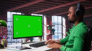 Black employee in his loft using green screen PC to analyze sales reports and plan business growth, remote job. Young man working from home doing project research, typing on computer. Camera B. video