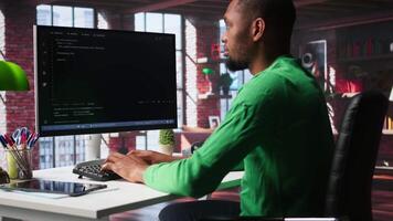 Black technician in home office looks at UI on computer monitor, typing and executing programming code. IT worker using coding application to write and test code in modern apartment. Camera B. video