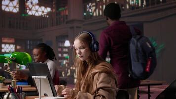 Young girl listening to educational documentary on laptop and taking notes, using modern non conventional way to study in the quietness of university library. Collects academic data. Camera B. video