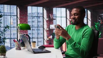 Man taking break from work to check his social media apps on mobile phone, having fun relaxing at home. Young guy in loft pausing tasks to use social network application on phone. Camera B. video