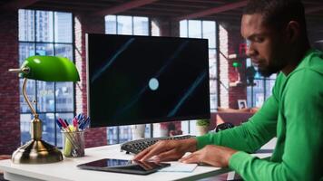 Black freelancer at home watching business conference on PC monitor, looking over statistics charts. Remote worker in apartment office follows presentation graphs on computer. Camera B. video