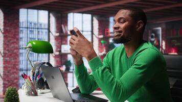 Man at home using smartphone to compose social media posts, producing content as a freelancer. African american guy monitoring profile engagement using mobile phone. Camera B. video