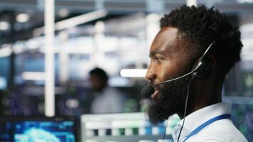 Data center employee talking in headset mic, coordinating technicians setting up hardware for artificial intelligence deployment. Server room worker engages in remote call with engineers, camera B video