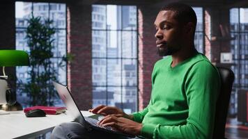 African american remote worker solving daily job assignments, finishing his tasks online from the comfort of his modern loft. Young adult teleworker connected on web, multitasking. Camera B. video