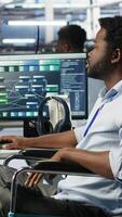 Vertical Data center engineer in wheelchair using AI requesting help from coworkers during security breach. Server farm worker with disability aided by colleagues to fight hacker attack stealing data, camera B video