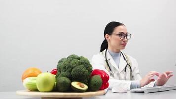 A nutritionist takes notes and works at a computer in a bright medical office. Fresh vegetables and fruits are on the table. The concept of healthy eating. Advertising video