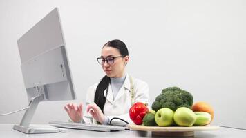 A nutritionist takes notes and works at a computer in a bright medical office. Fresh vegetables and fruits are on the table. The concept of healthy eating. White background, free copy space video