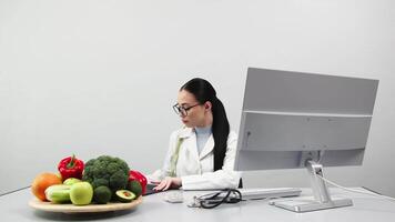 A nutritionist takes notes and works at a computer in a bright medical office. Fresh vegetables and fruits are on the table. The concept of healthy eating video