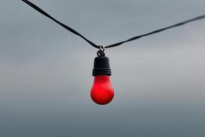 Glowing red light bulb hanging against a cloudy sky illuminating the dark atmosphere photo
