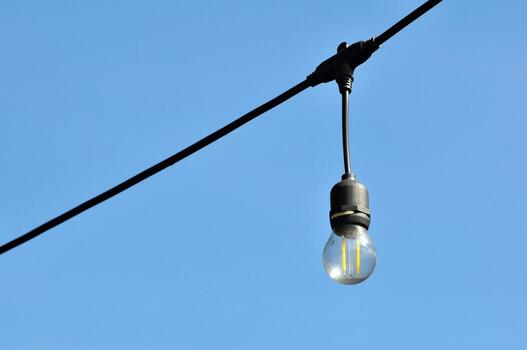 Single LED light bulb hanging from an overhead cable with a clear blue sky in the background photo