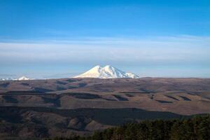 The snow-capped peak of Elbrus rises above the hills against the backdrop of a clear sky. photo