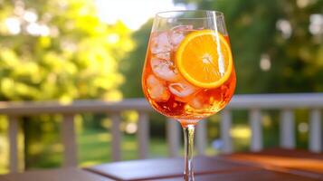 Aperol Spritz with ice and orange slice in a wine glass on sunlit terrace table with garden backdrop photo