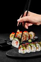Woman using chopsticks picking sushi rolls from plate on black background photo
