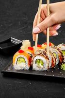 Woman picking up sushi rolls from plate with chopsticks on dark background photo