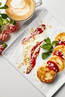 Delicious syrniki with sour cream, raspberry jam, almond flakes, and mint served with cappuccino and roses photo
