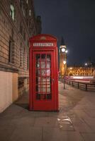 A red telephone booth stands on a quiet London street at night, with Big Ben illuminated in the background. Streetlights and motion blur add dynamic elements. photo