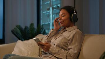 Peaceful relaxed African American woman ethnic girl in headphones listening music relaxing on cozy sofa enjoy song with mobile phone listen audio in earphones rest lazy weekend at night evening home video