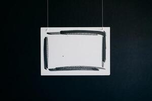 A rectangular white canvas with black painted edges hanging against a dark blue background wall photo
