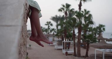 Tourist legs dangling freely while sitting on wall, overlooking tropical resort landscape with swaying palm trees and colorful beach umbrellas surrounding scenic background video