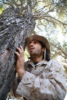 Scared Soldier Hides Behind The Trunk Of A Tree In The Mountains From Enemy photo