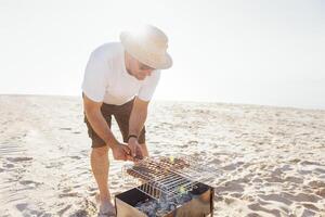 A young man in a summer casual outfit is having a barbecue on the sandy beach. A male in sunglasses and a straw hat is frying sausages on the seashore. photo