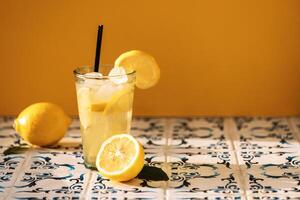 Close-up of a glass of delicious lemonade with ice cubes on a table made of white tiles with blue patterns. A cup of cool citrus fruit. A tasty sour drink with lemons. photo