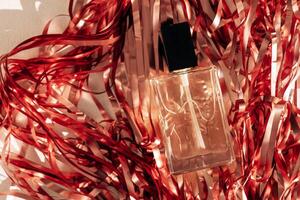 Close-up of a transparent glass perfume bottle with a black lid on a shiny red background made of metallized paper filler. A vial of fragrant water on foil shredded paper. photo