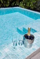 Two glasses of empty glasses and ice bucket with champagne on the side of the pool. A chilled bottle of fragrant sparkling wine on vacation. Clear blue water and bright sunlight. photo