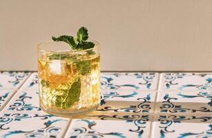 Close-up of a glass of mineral water with lemon and fresh mint and ice cubes on a table made of white tiles with blue patterns. A cup of cool, delicious, healthy drink. photo