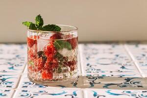 Close-up of a glass of mineral water with raspberries and fresh mint and ice cubes on a table made of white tiles with blue patterns. A cup of cool, delicious, healthy drink. photo