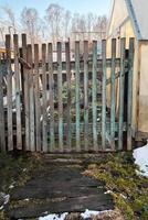 Rustic wooden garden gate leading to a backyard area during early spring. The scene captures remnants of snow melting on the ground, with bare trees in the background photo
