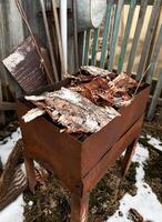 Rusty metal grill filled with dry birch bark, ready for lighting a barbecue fire. The setup is outdoors, surrounded by rustic materials photo