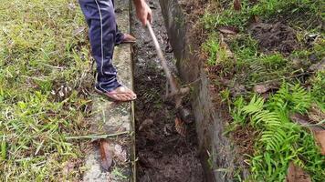 An Indonesian man cleans a dirty water channel on the side of a village road using a hoe. Community sanitation efforts in rural areas video