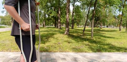 Impersonal man walking with crutches in a busy park showing determination and resilience against the background of nature, copy space, banner photo