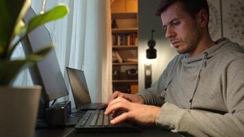 Man sitting by table and looking at computer monitor. Concentrated freelancer using desktop and laptop by window. Remote work from home office video