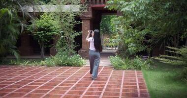 Handheld back view shot, Solo female tourist enjoys taking photos with camera while taking a leisurely stroll amidst serene surroundings and the cultural richness of the place. video